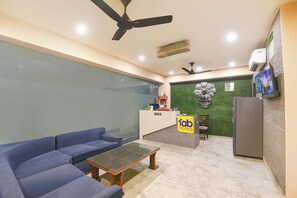 Ein Hotel-Rezeptionsbereich mit Empfangstresen, einem blauen Sofa, einem Couchtisch, einem Deckenventilator und einem an der Wand montierten Fernseher.