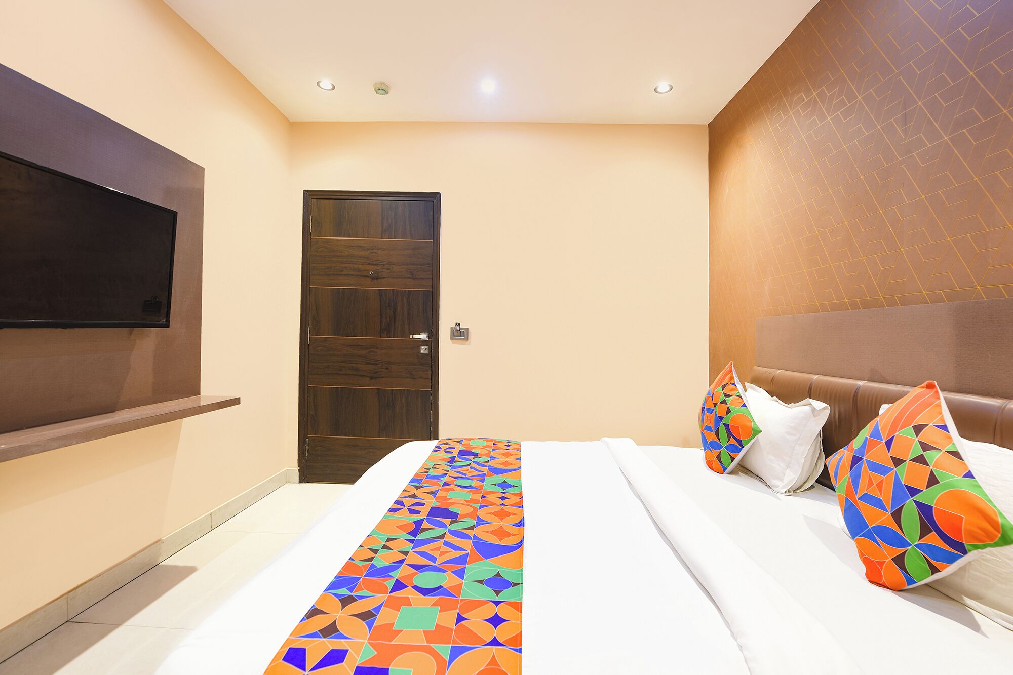 A hotel room with a bed, a TV mounted on the wall, a door, and a patterned runner.