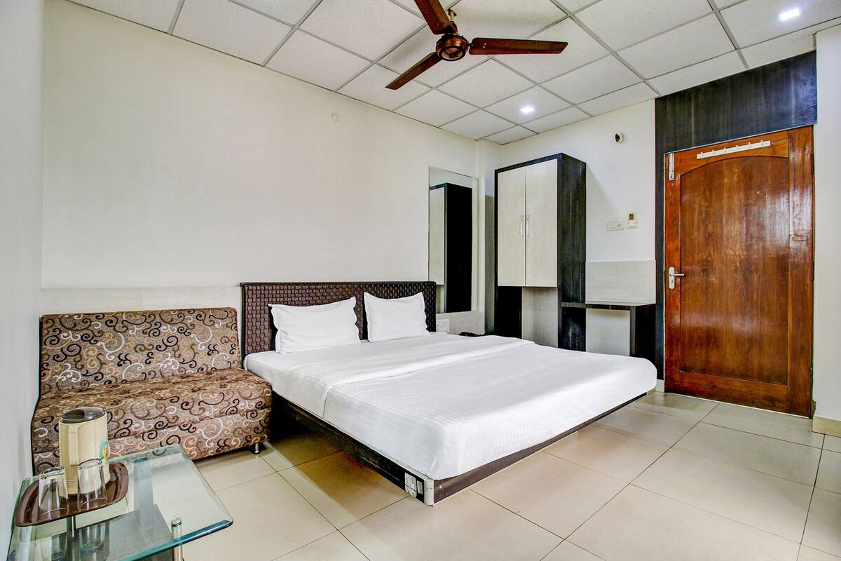 A hotel room with a bed, a sofa, a glass table with a tray, a ceiling fan, and a wooden door.