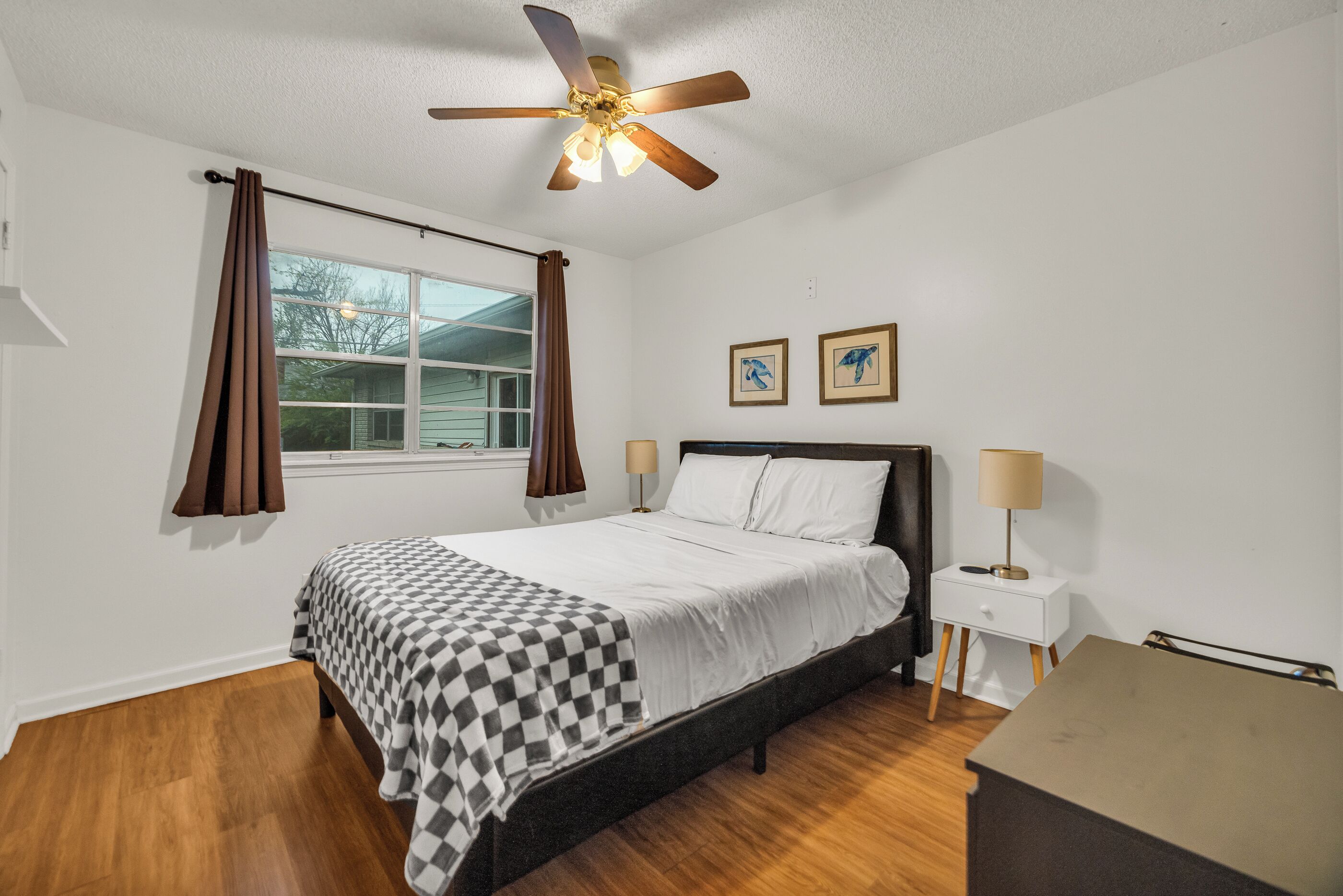 A bedroom with a bed, a ceiling fan, a window with curtains, two wall-mounted lamps, and a small bedside table.