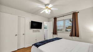 Une chambre à coucher avec un lit, un ventilateur de plafond, une télévision et une fenêtre avec des rideaux.