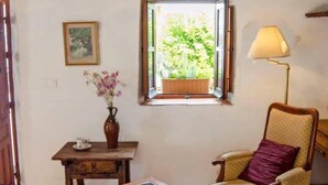 Ein gemütliches Zimmer mit einem Holztisch, einem Sessel, einem Fenster mit Blick ins Grüne, einer Lampe und einem kleinen Tisch mit Blumen in einer Vase.