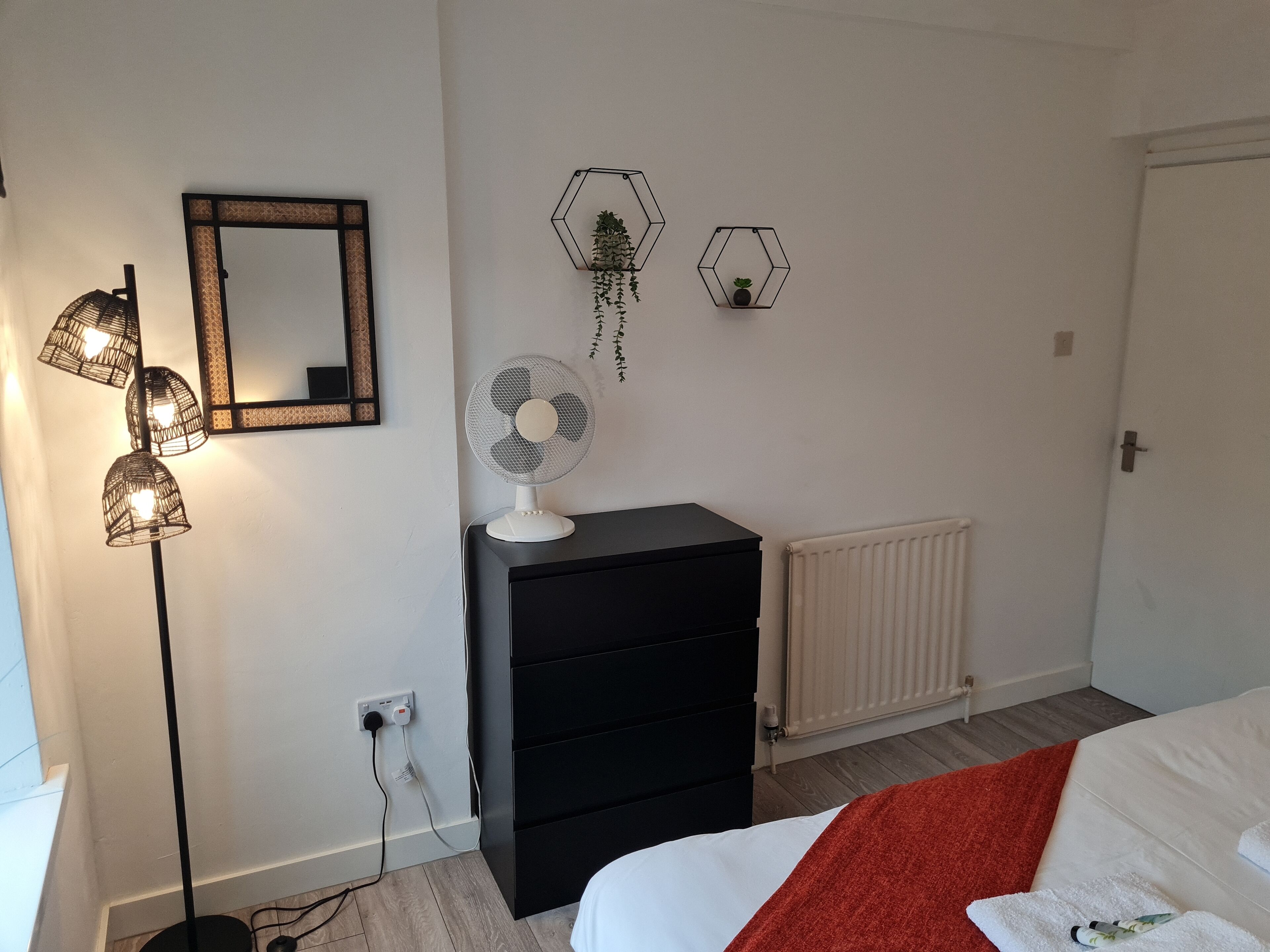 A bedroom with a bed, a dresser, a standing fan, a mirror, and a radiator.