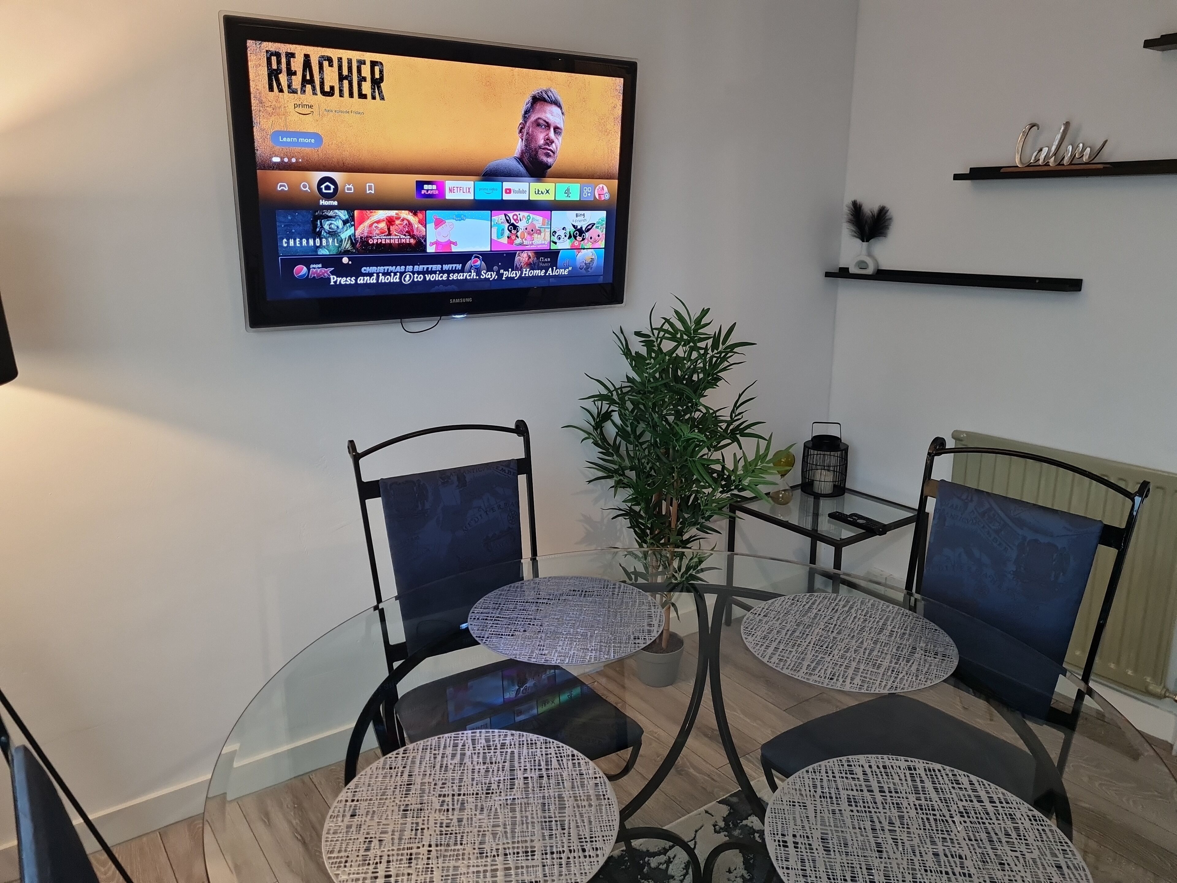 Una sala de estar moderna con una mesa de comedor de vidrio rodeada de sillas, un televisor montado en la pared y una pequeña planta en maceta.