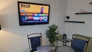 Ein modernes Wohnzimmer mit einem Glastisch, umgeben von Stühlen, einem an der Wand montierten Fernseher und einem kleinen Topfpflanzen.