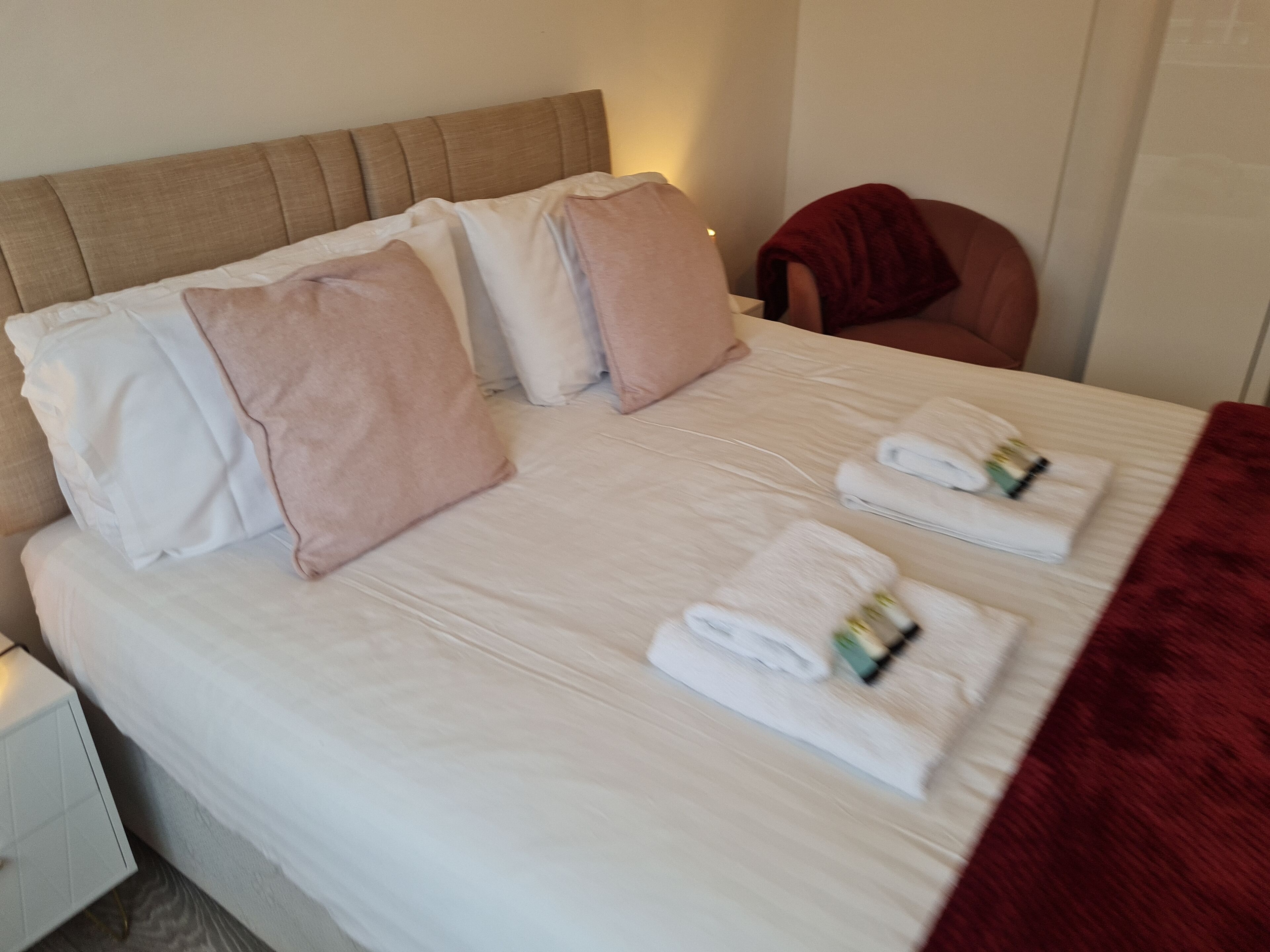 Cama bem arrumada com lençóis brancos, almofadas rosa e brancas, e toalhas enroladas.