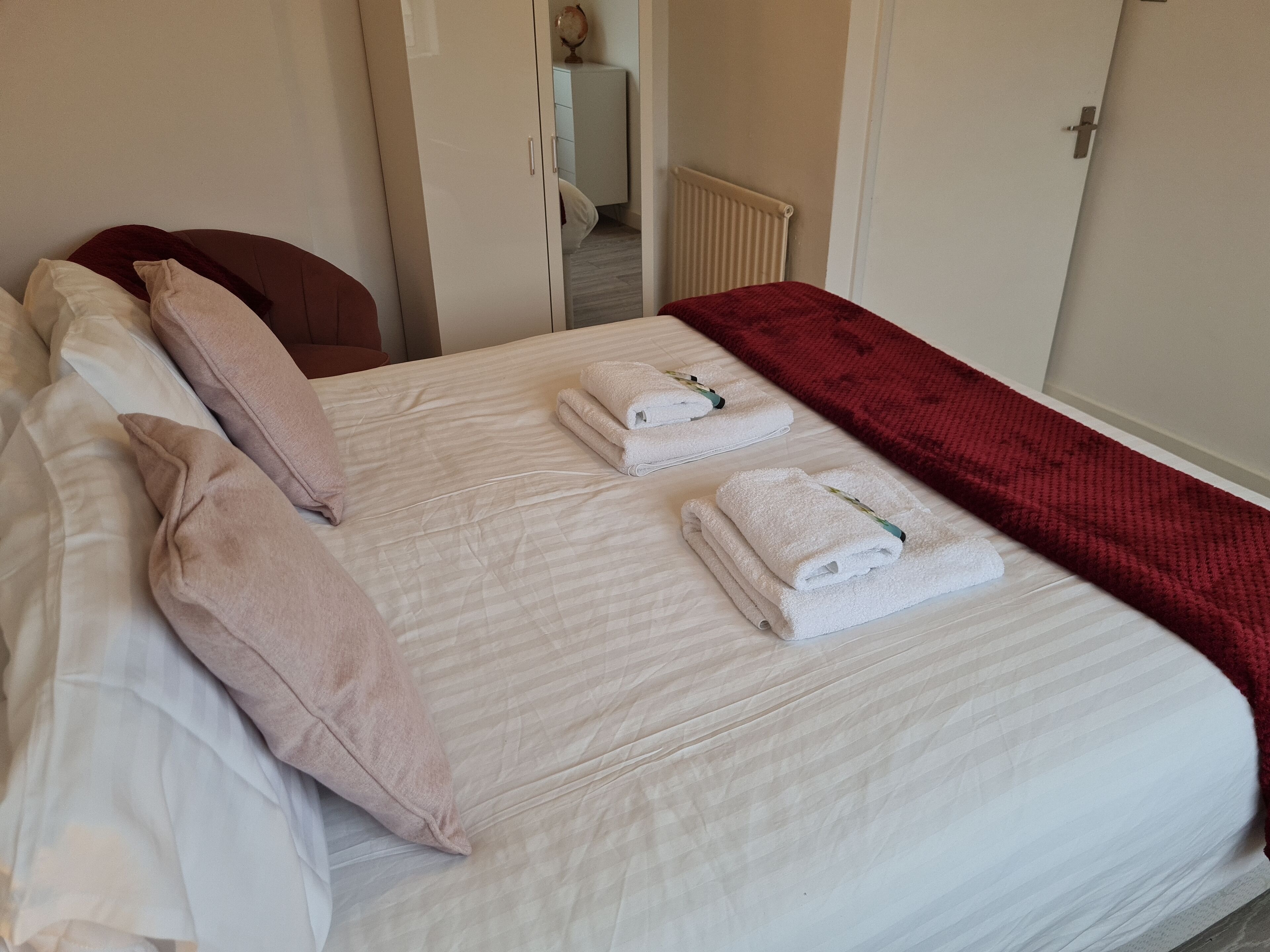 Una cama bien hecha con sábanas blancas, un plaid rojo y toallas dobladas.