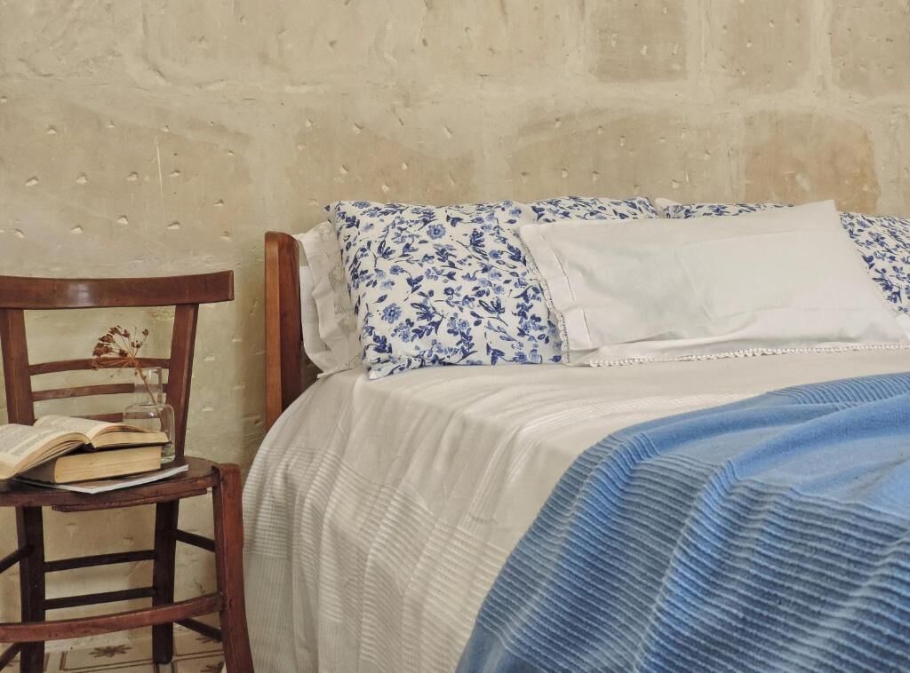 Ein Bett mit weißblauer Blumendecke, einem weißen Kissen und einem Holzstuhl mit Buch und Glas.
