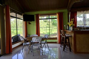 Une pièce comprenant une table à manger, des chaises, un téléviseur fixé au mur et une fenêtre avec des rideaux.