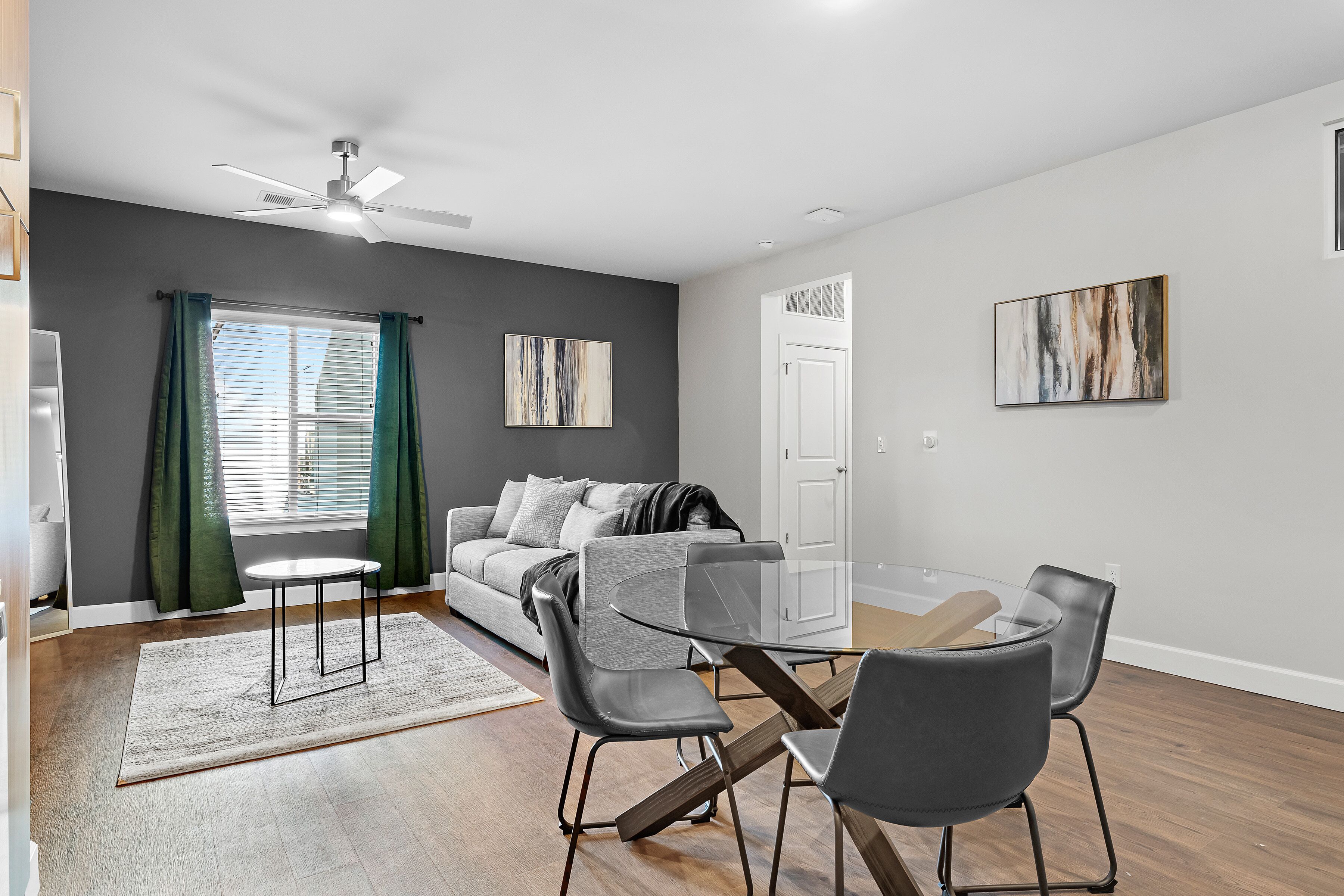 A modern living room with a glass dining table, grey chairs, a grey sofa, and a ceiling fan.