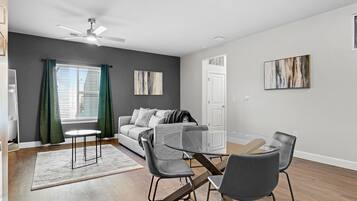 A modern living room with a glass dining table, grey chairs, a grey sofa, and a ceiling fan.