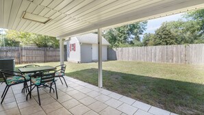 Ein überdachter Patio-Bereich mit Tisch und Stühlen, eine kleine Gartenhütte und ein Holzzaun im Hintergrund.