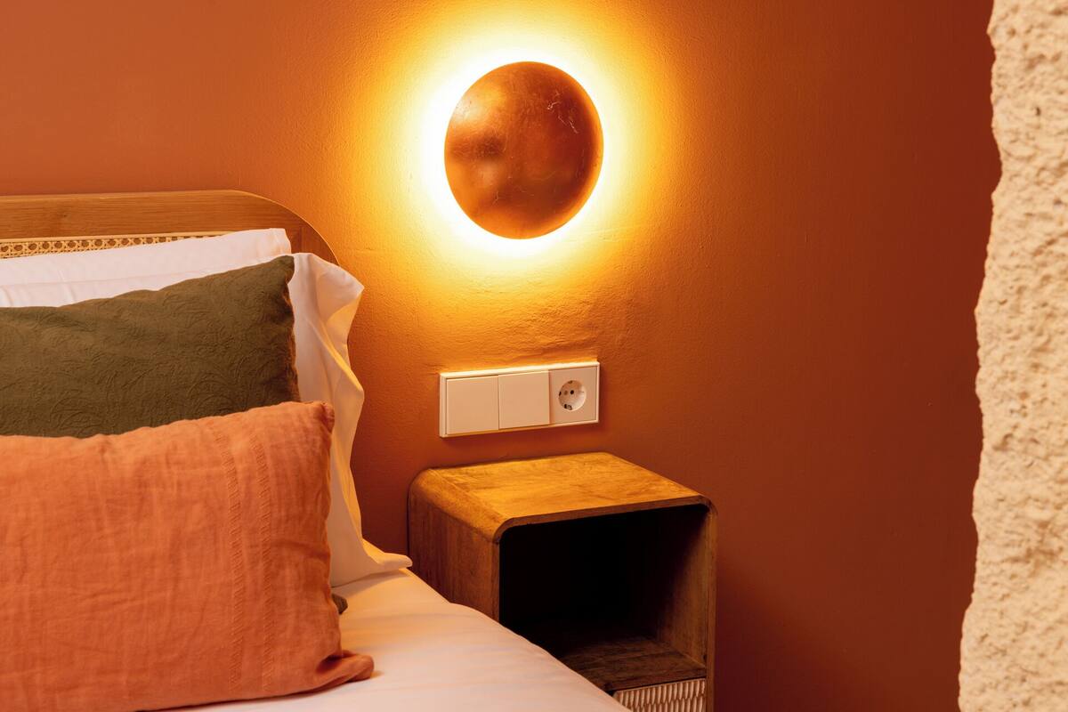 Un dormitorio con cama, almohadas, una mesita de noche y una lámpara de pared.