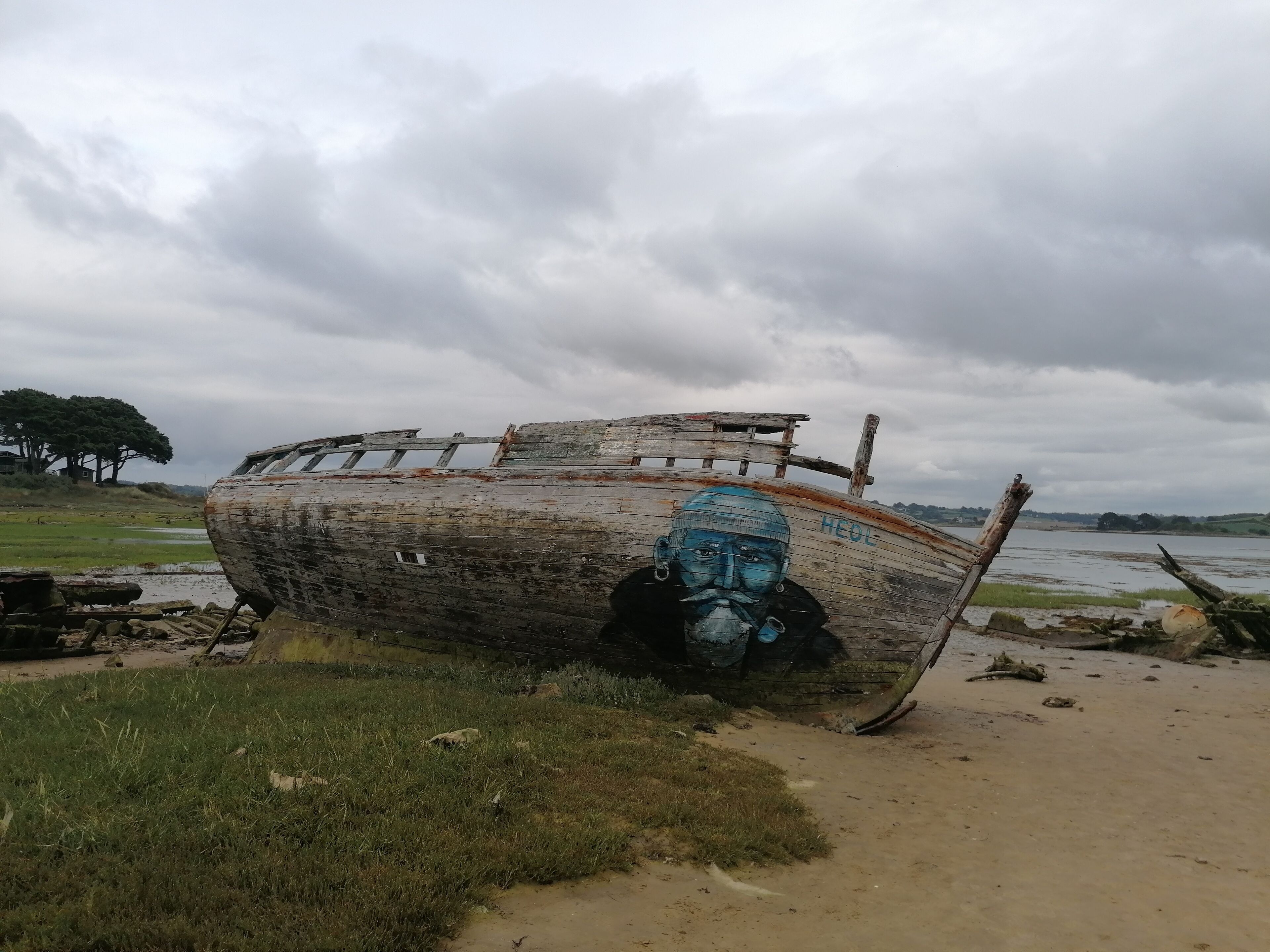 An old, weathered boat with graffiti on its side, resting on a grassy area near a body of water.