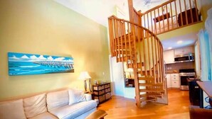 A living room with a wooden staircase, a sofa, a painting, and a kitchen in the background. - Charming 1BR Cottage : Steps from Hemingway House & Old Town (Key West)