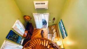 A room with a spiral staircase, a sofa, a small table, a plant, and a wall-mounted air conditioner. - Charming 1BR Cottage : Steps from Hemingway House & Old Town (Key West)