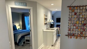 A modern room with a bed, a small table, and a kitchenette. There is a sign above the door that reads "NEW SEABURY". -   EXPLORE NEW SEABURY from our 2/2 Townhome - views + beach+ pool+ fireplace.  (Mashpee)