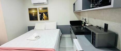 A hotel room with a bed, a small kitchenette, and a wall-mounted air conditioner.