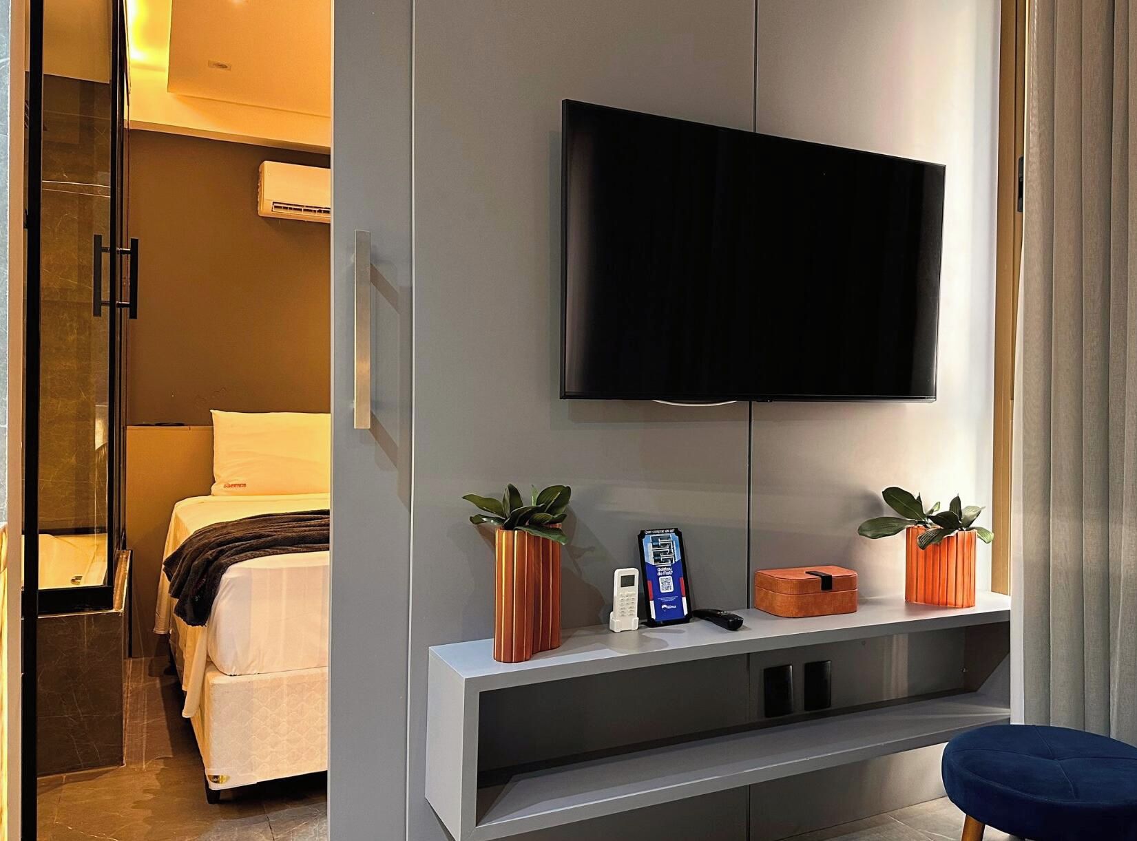 Habitación de hotel con cama, televisión montada en la pared, mesita de noche con teléfono y una caja pequeña, y ventana con cortinas.