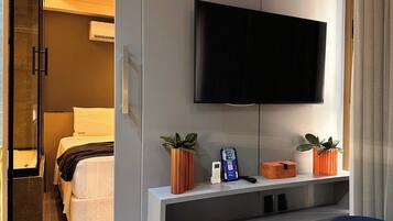 Habitación de hotel con cama, televisión montada en la pared, mesita de noche con teléfono y una caja pequeña, y ventana con cortinas.