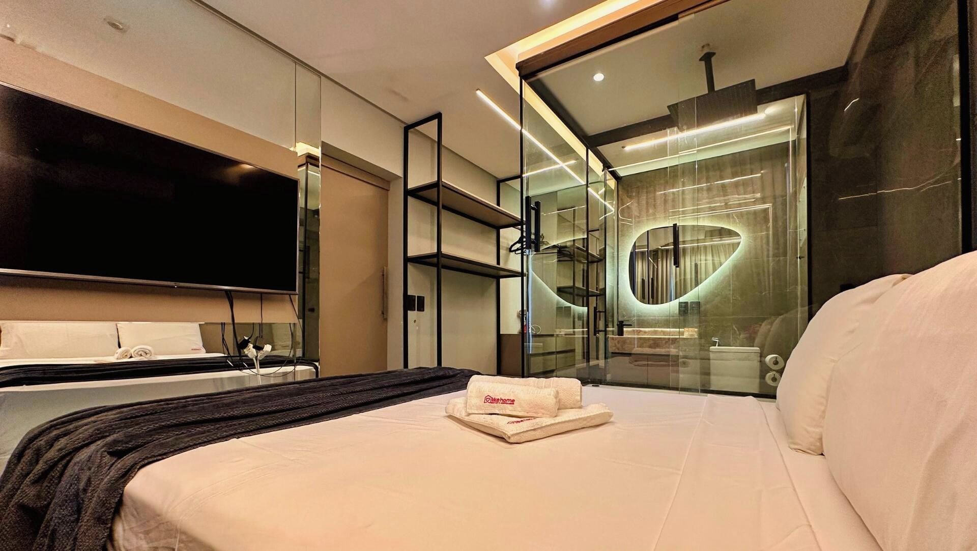 A modern hotel room with a large bed, a flat-screen TV, a glass-enclosed shower area, and a minimalist design.