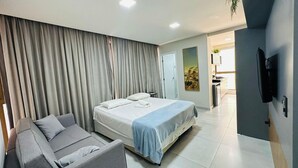 Un dormitorio moderno con una cama, un sofá, un televisor y un baño visible al fondo.