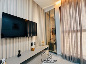 A modern living room with a flat-screen TV mounted on a wall with vertical blinds, a minimalist console table with decorative items, and a large mirror reflecting the room. - Co-House (João Pessoa)