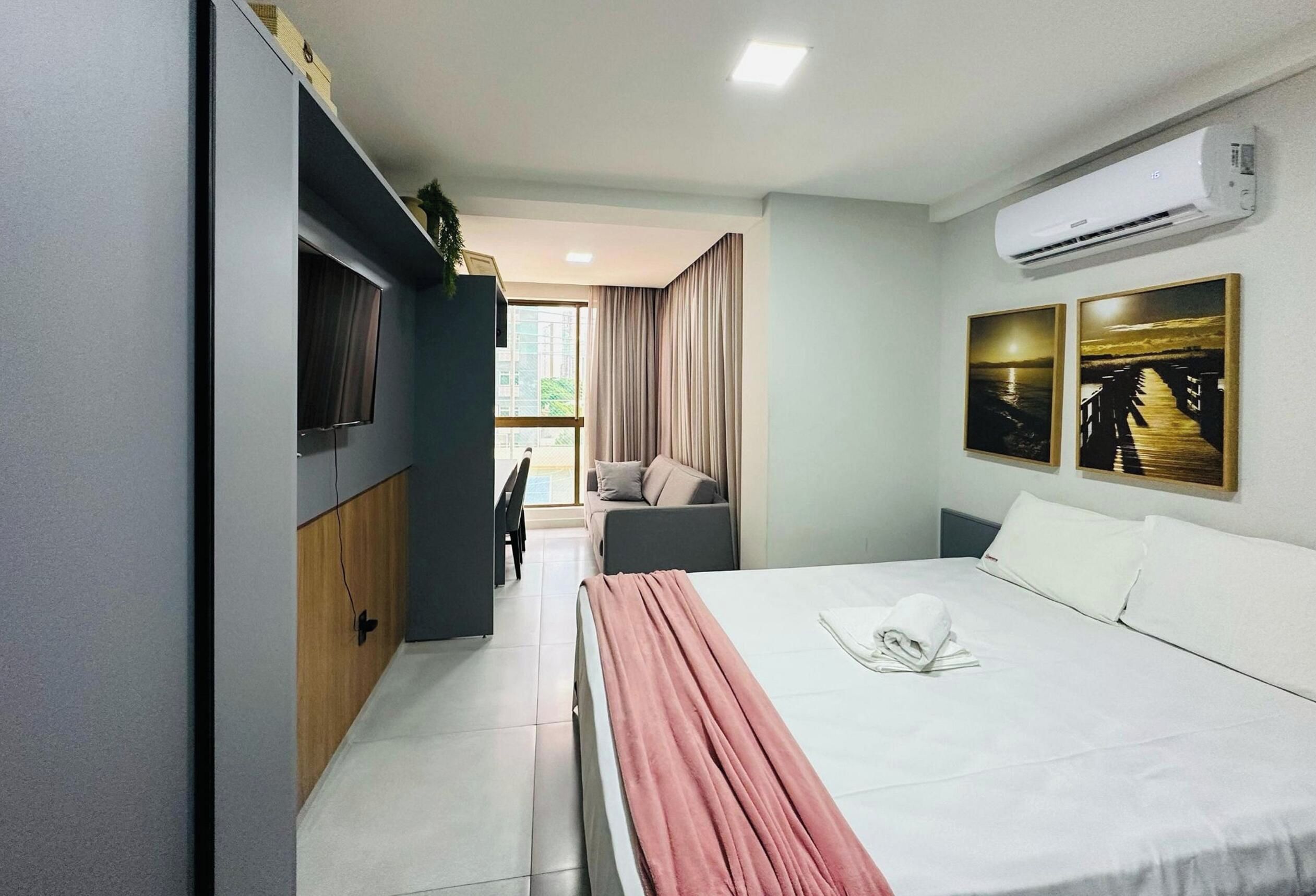 A hotel room with a bed, a desk, a chair, an air conditioning unit, and two paintings on the wall.