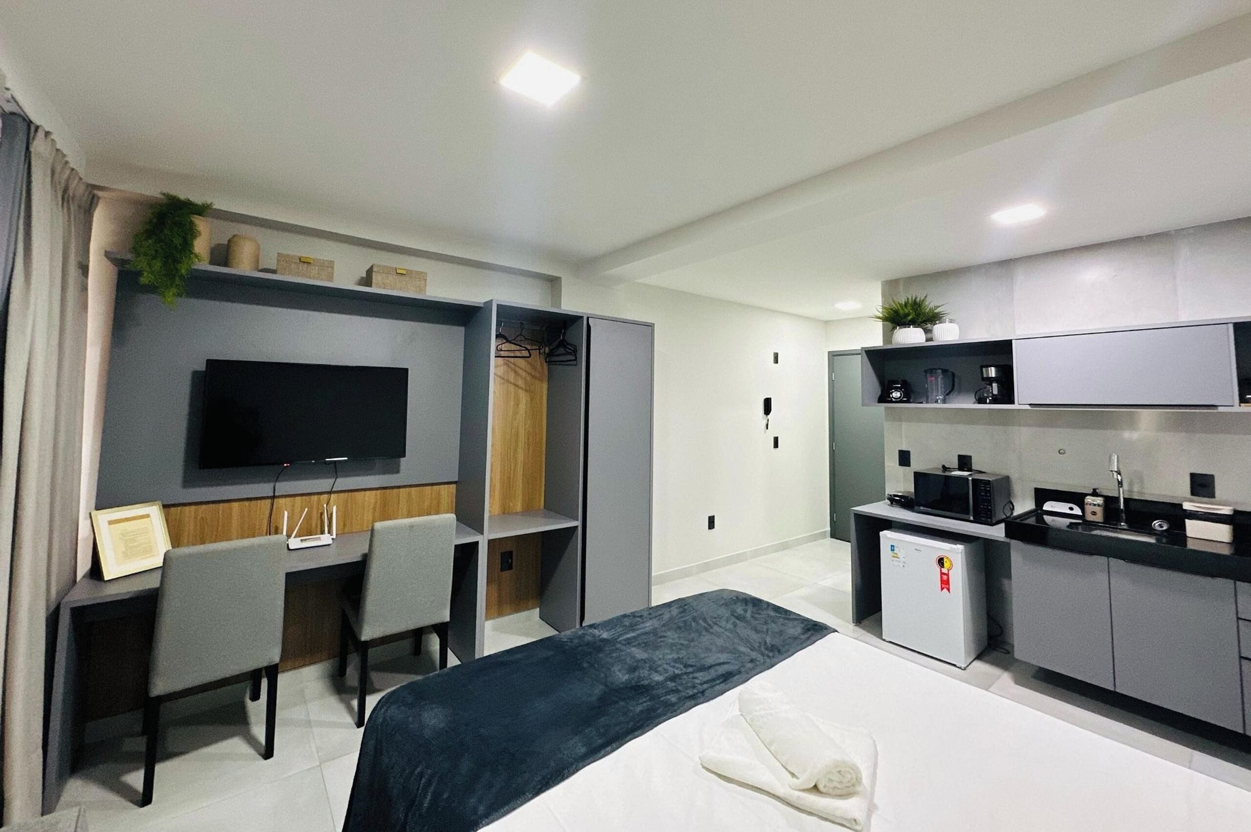 A modern hotel room with a bed, a desk, a TV, a kitchenette, and a wardrobe.
