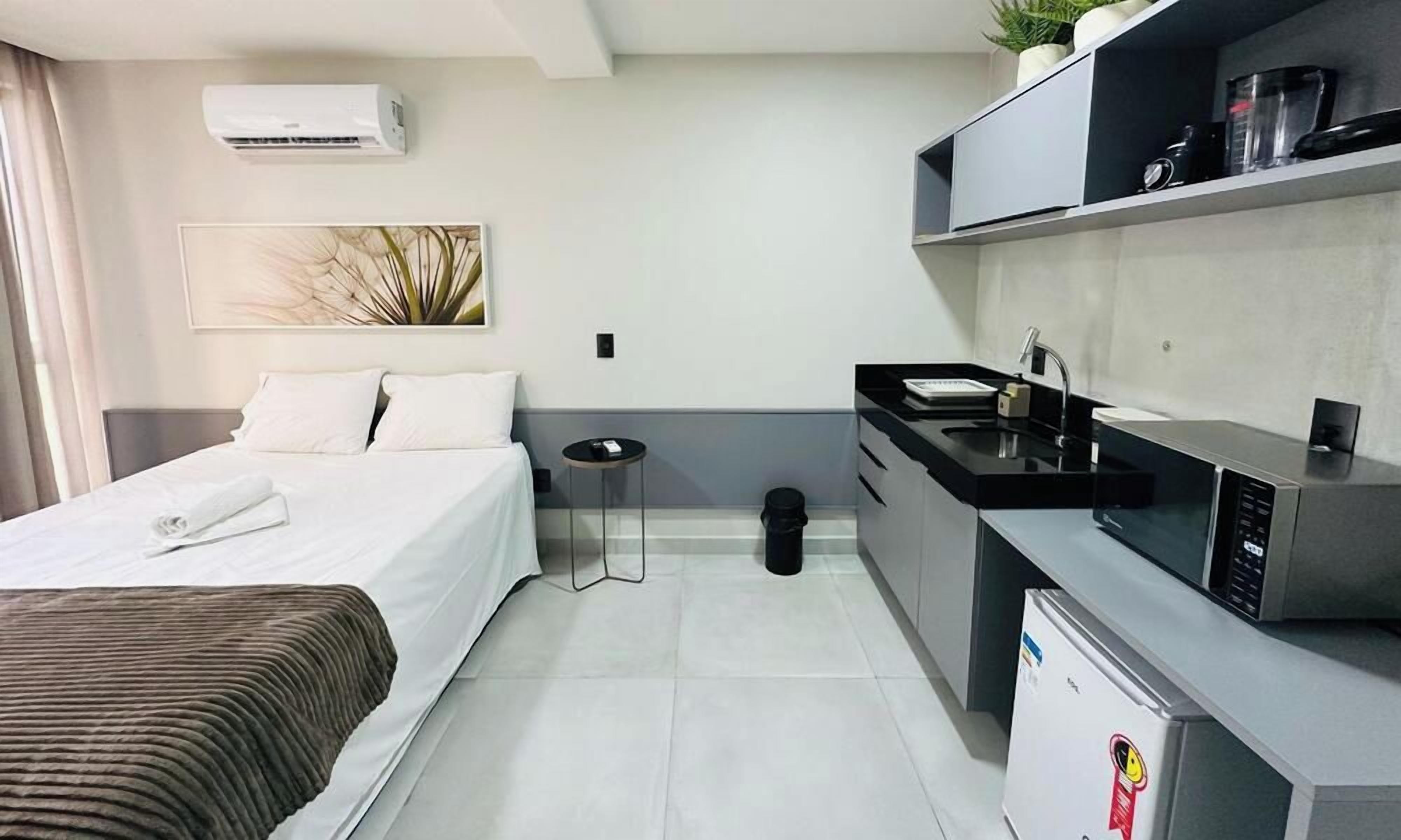 A compact hotel room with a bed, a small kitchenette, a trash bin, and a wall-mounted air conditioner.