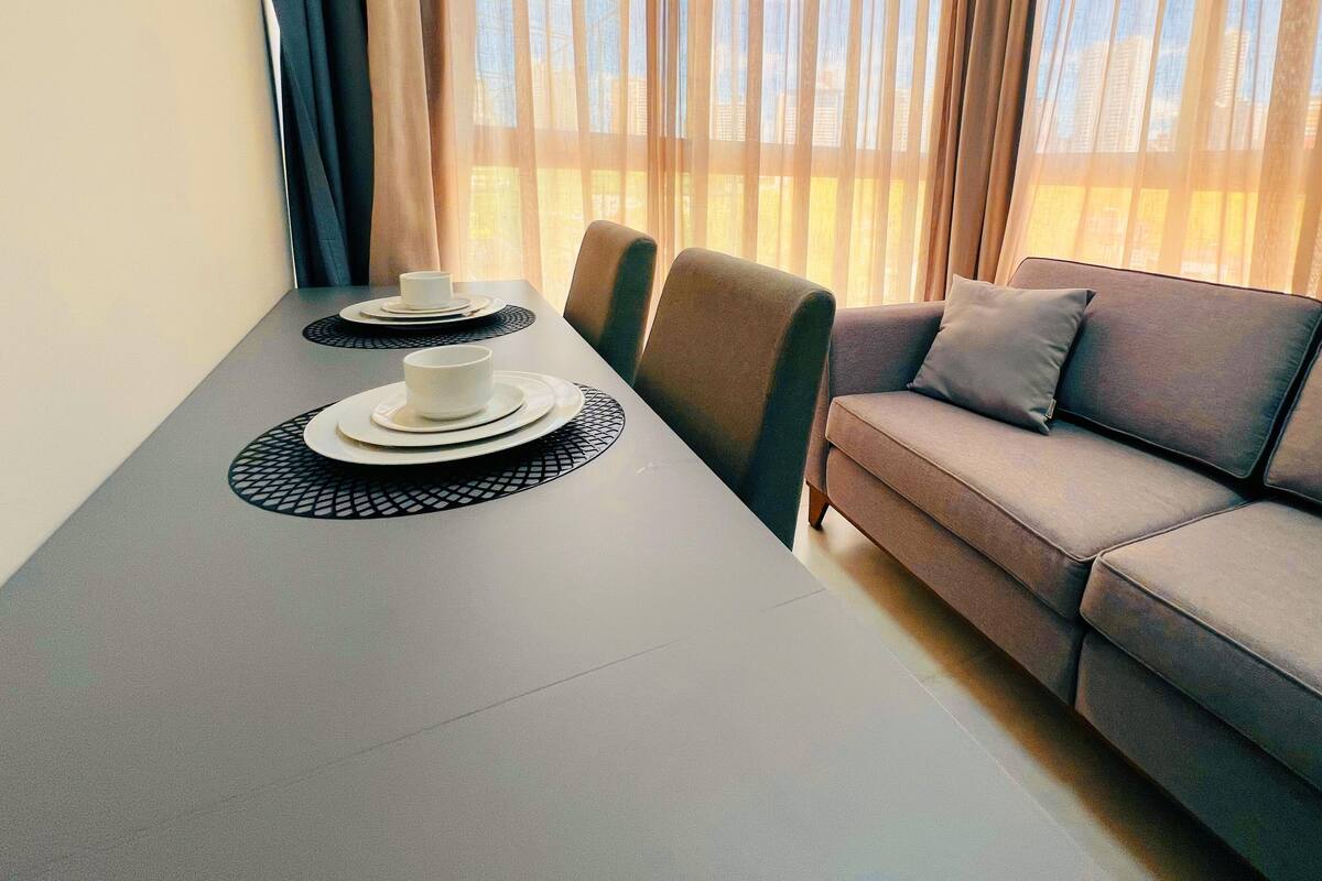 Una habitación moderna con una mesa de comedor dispuesta para dos personas, un sofá gris y vistas a través de cortinas translúcidas.