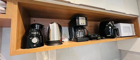 Una cocina con un estante de madera que contiene una cafetera, un hervidor, un microondas y un tostador.