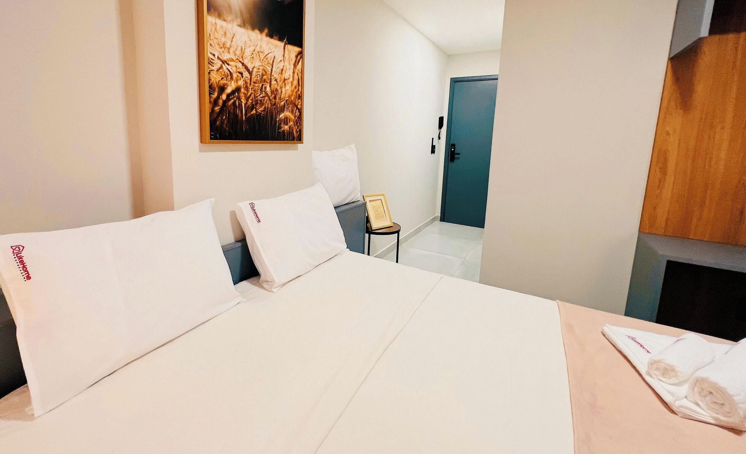 Una camera d'albergo con un letto, cuscini, un tavolino, una sedia, una porta e un quadro appeso al muro.