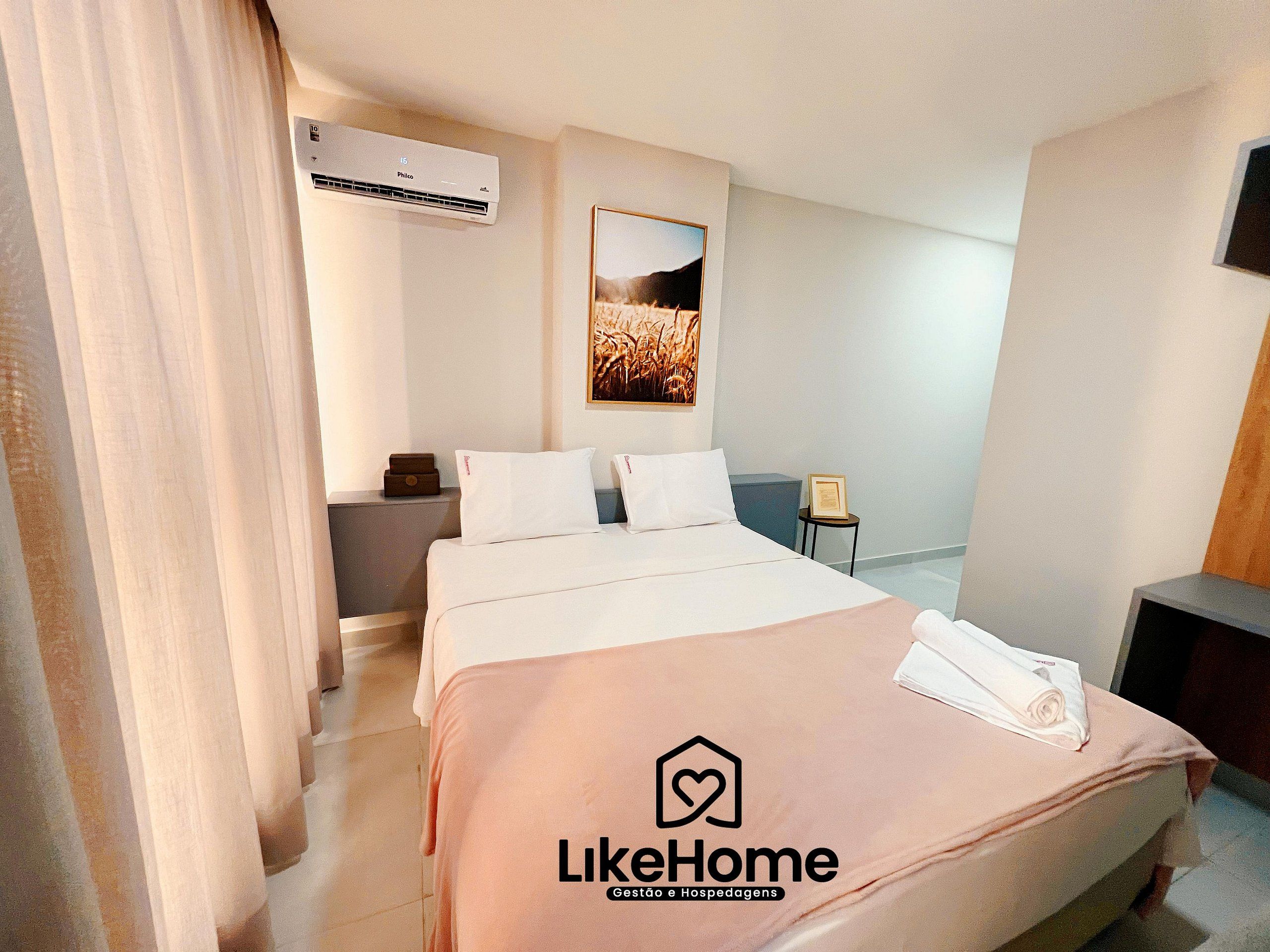 A hotel room with a bed, a bedside table, an air conditioning unit, a framed picture on the wall, and a logo on the bedspread.
