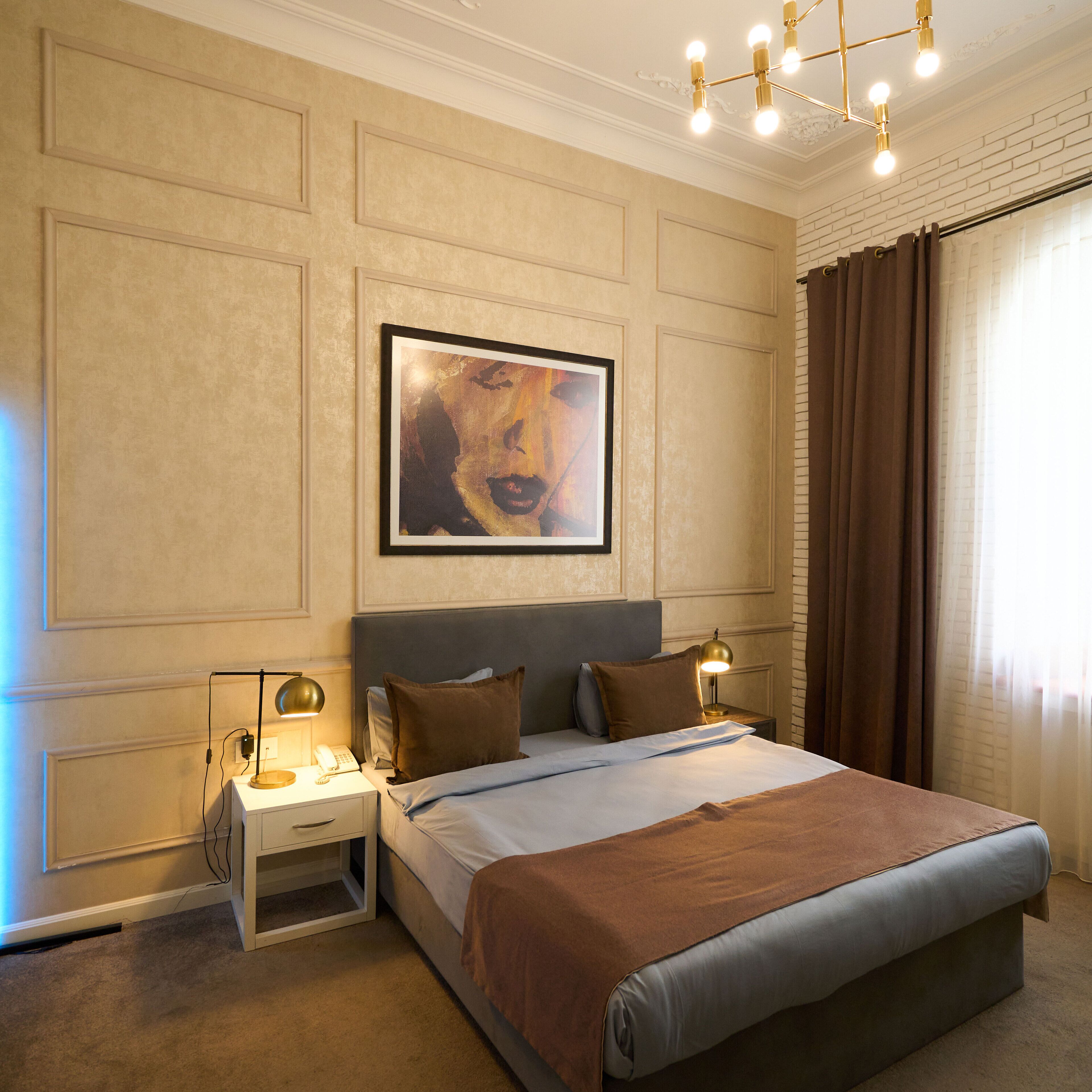 A hotel room with a large bed, two bedside tables, a nightstand, a framed artwork on the wall, and a window with curtains.