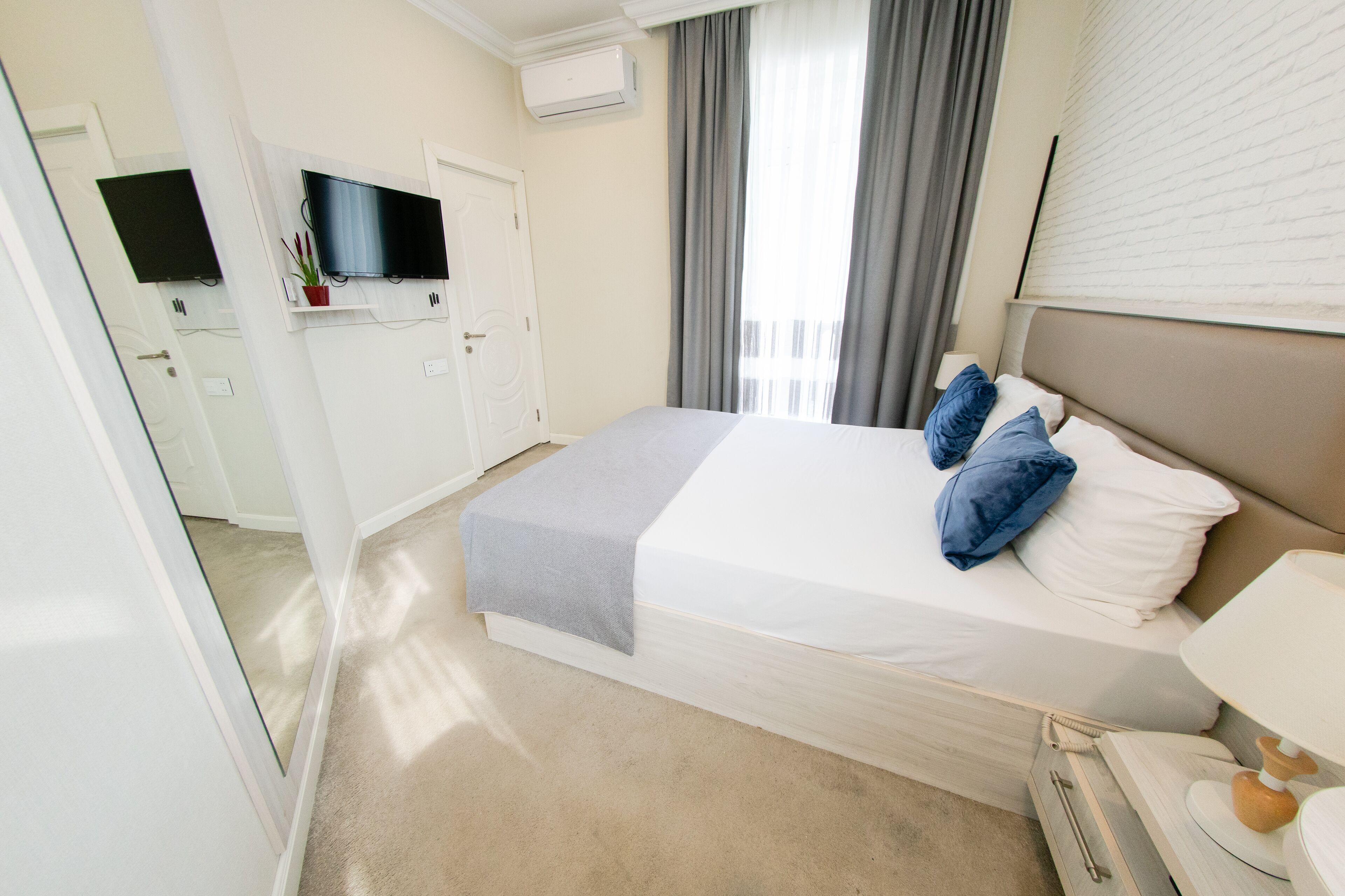 A hotel room with a bed, a TV mounted on the wall, a window with curtains, and a bedside table with a lamp.