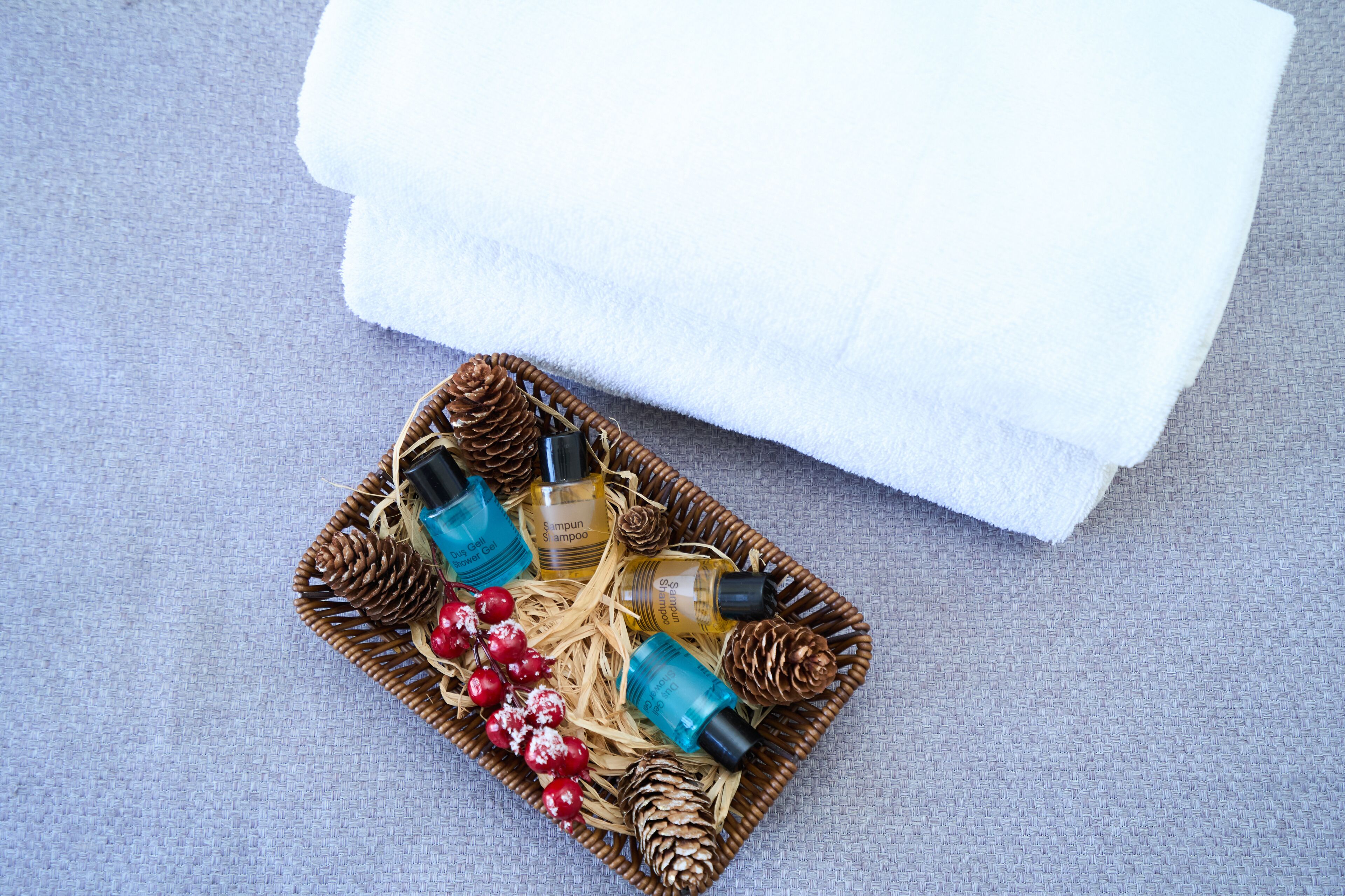 A wicker basket with shampoo bottles, pine cones, and red berries on a grey fabric background.