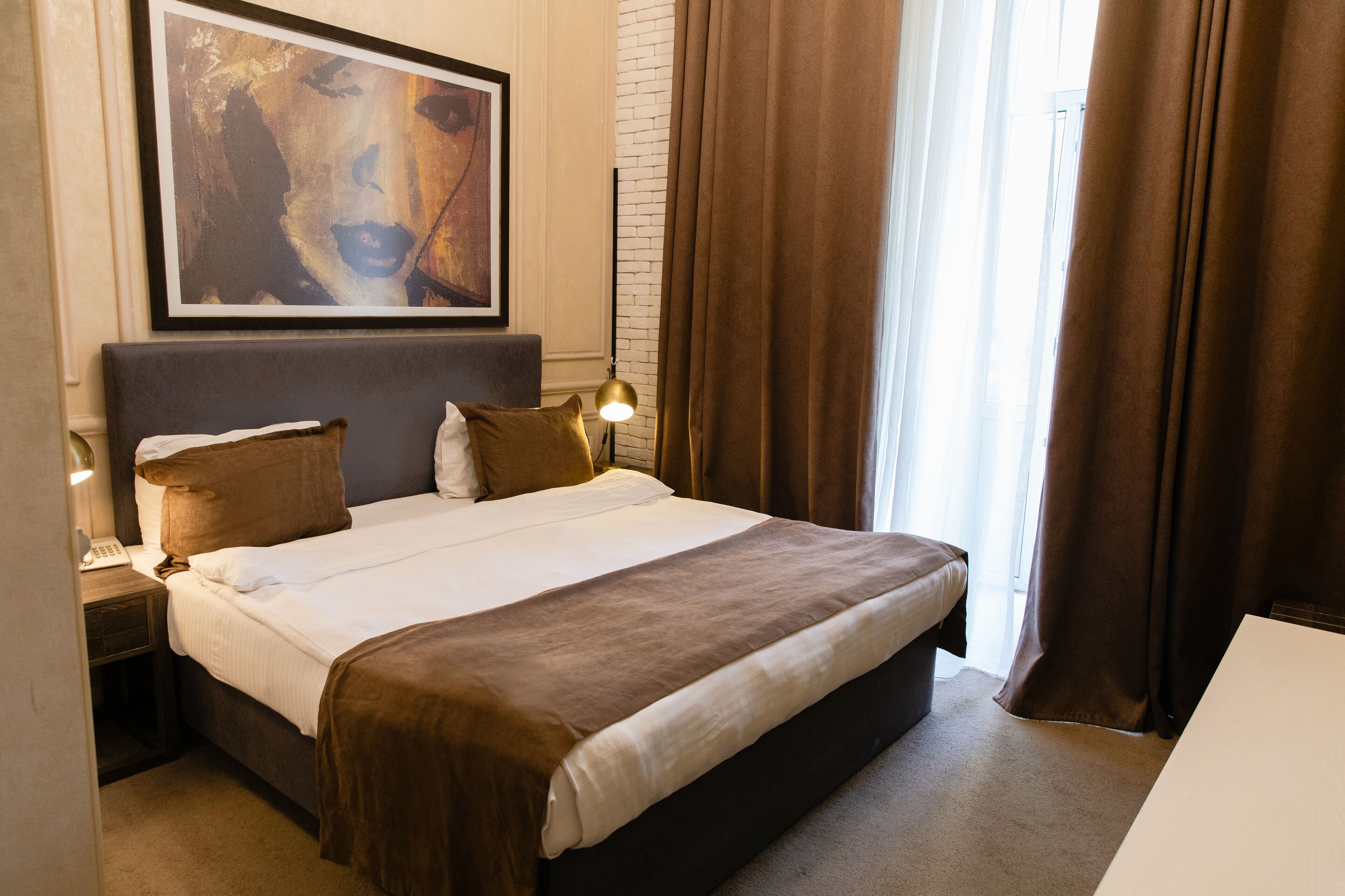 Habitación de hotel con una cama grande, una mesita de noche con lámpara, un cuadro en la pared y una ventana con cortinas.