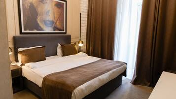 Habitación de hotel con una cama grande, una mesita de noche con lámpara, un cuadro en la pared y una ventana con cortinas.