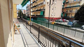 A balcony with a view of a multi-story building with balconies, a street with parked cars, and a green fence. - Renesmee Beach Apartment a 150 Metri Dalla Spiaggia, con Posto Auto Gratuito (Pietra Ligure)