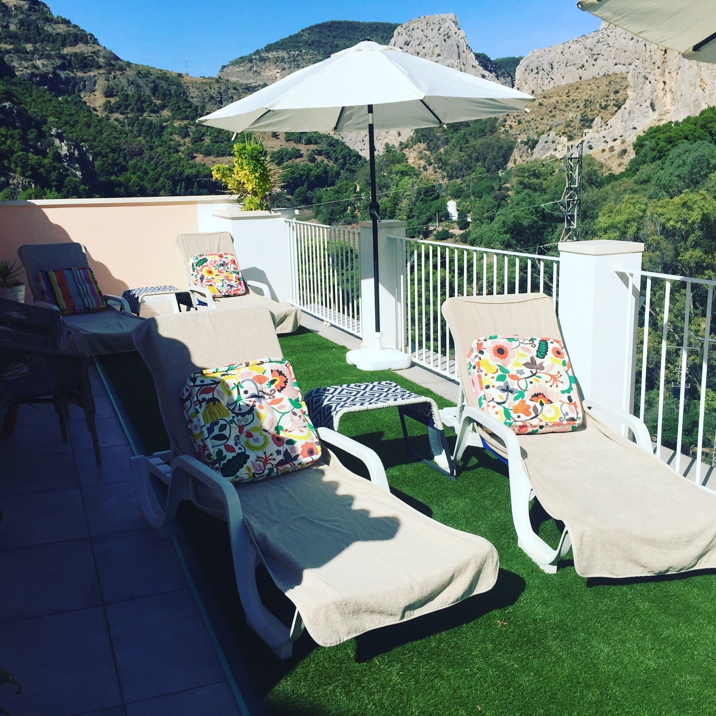 Una terraza en la azotea con dos sillones de descanso, una mesita y una sombrilla grande. La terraza ofrece vistas a colinas verdes y montañas rocosas al fondo.