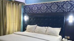 Un dormitorio con una cama grande, un cabecero azul oscuro, un papel tapiz con diseño, un equipo de aire acondicionado y una mesita de noche con un teléfono.