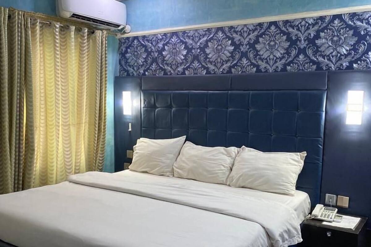 Un dormitorio con una cama grande, un cabecero azul oscuro, un papel tapiz con diseño, un equipo de aire acondicionado y una mesita de noche con un teléfono.