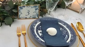 Una mesa dispuesta con un plato azul y blanco, cubiertos dorados y una servilleta blanca doblada sobre el plato.