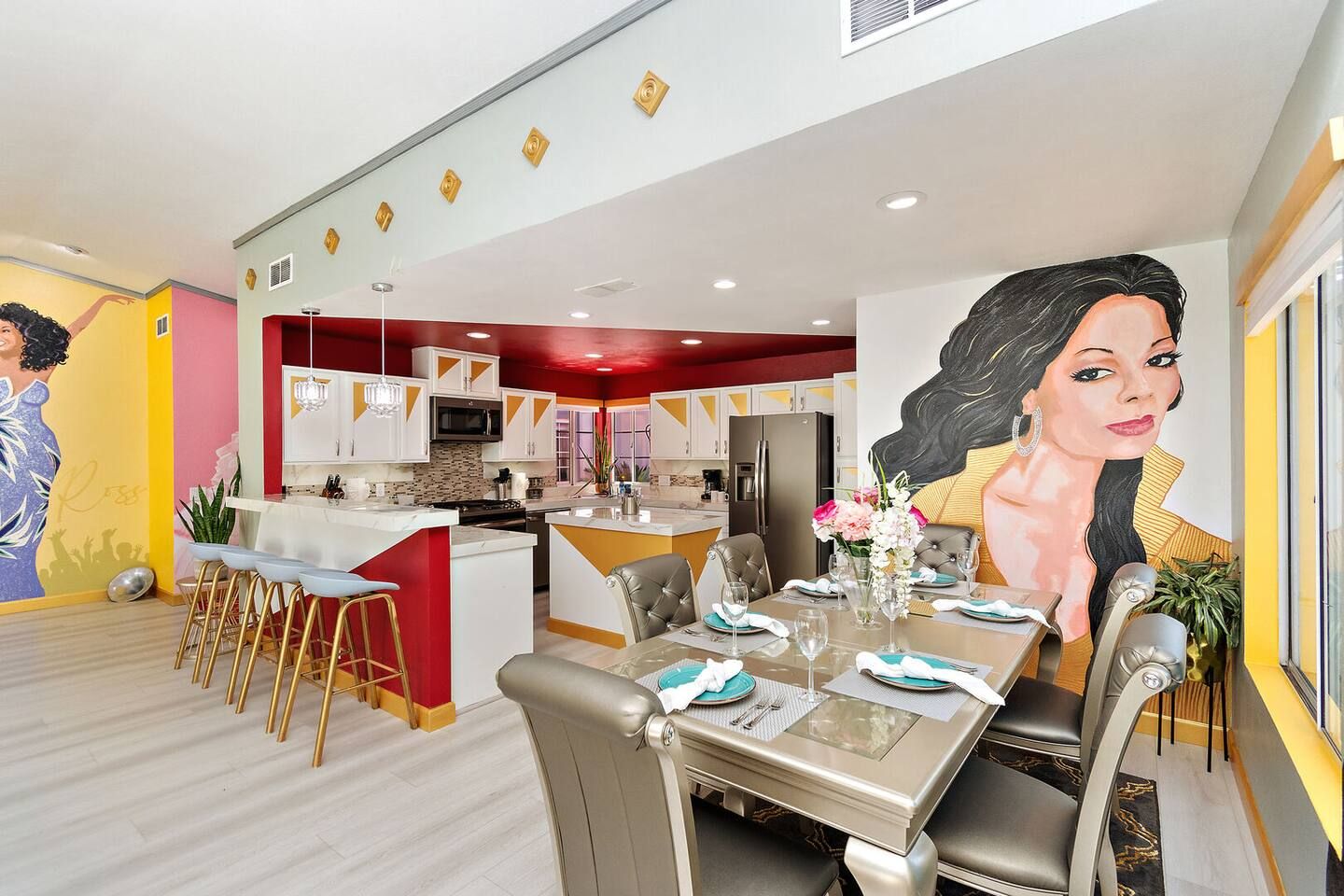 A modern kitchen with a central island, dining area with a table set for four, and a large mural on the wall.