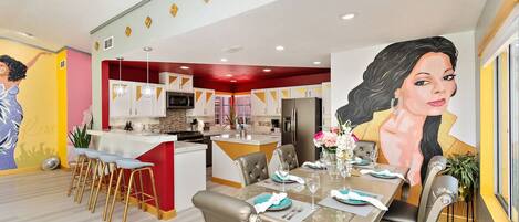 A modern kitchen with a central island, dining area with a table set for four, and a large mural on the wall.