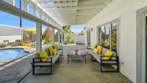 A covered outdoor area with a pool, two sofas with yellow cushions, and a table with a vase of flowers. - Rock N Royalty: Pool, Spa, Fire Pit, Pool Table! (Bermuda Dunes)