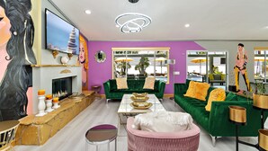 A modern living room with a fireplace, green sofas, a pink armchair, and a large TV. - Rock N Royalty: Pool, Spa, Fire Pit, Pool Table! (Bermuda Dunes)