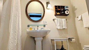 Un baño con lavamanos pedestal, un espejo, un perchero y un estante con artículos de aseo.