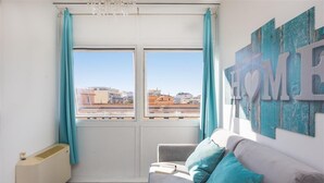 Une pièce avec un canapé, un radiateur et une vue sur des immeubles, visible à travers une fenêtre ornée de rideaux turquoise.